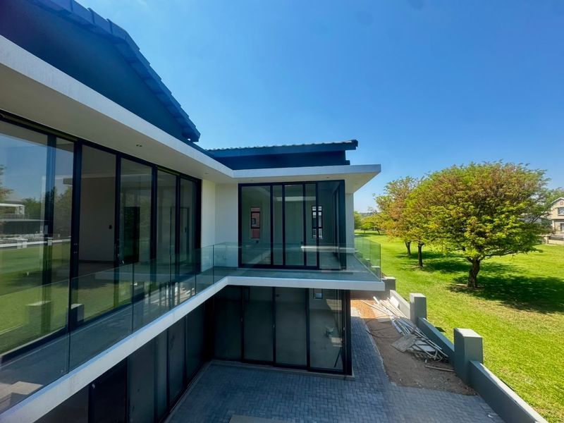 Rear elevation of a modern double-story home featuring extensive glass sliding doors, a glass balustrade balcony, and a paved patio area overlooking a green lawn.
