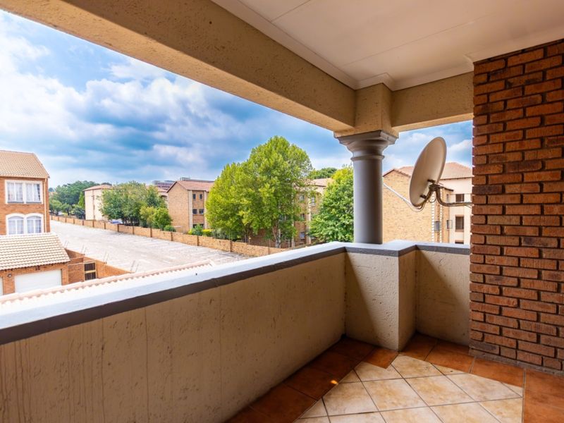 A covered balcony featuring tiled flooring, a brick accent wall, and a concrete column, offering an elevated view of the surrounding residential complex.