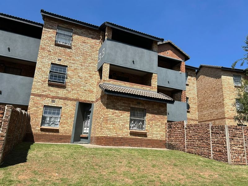 A multi-story apartment building featuring a face brick facade, tiled roofing, and private balconies with security bars, set against a clear blue sky.