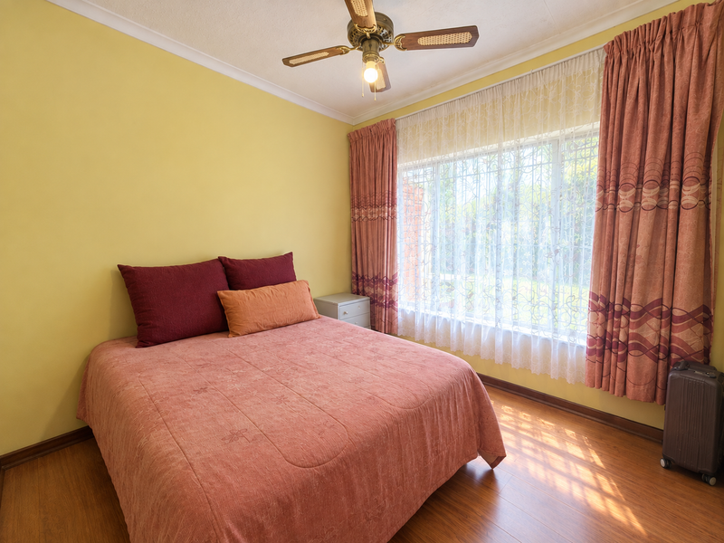 A well-lit bedroom featuring laminate flooring, a ceiling fan, and large windows with curtains, offering a comfortable and functional living space.