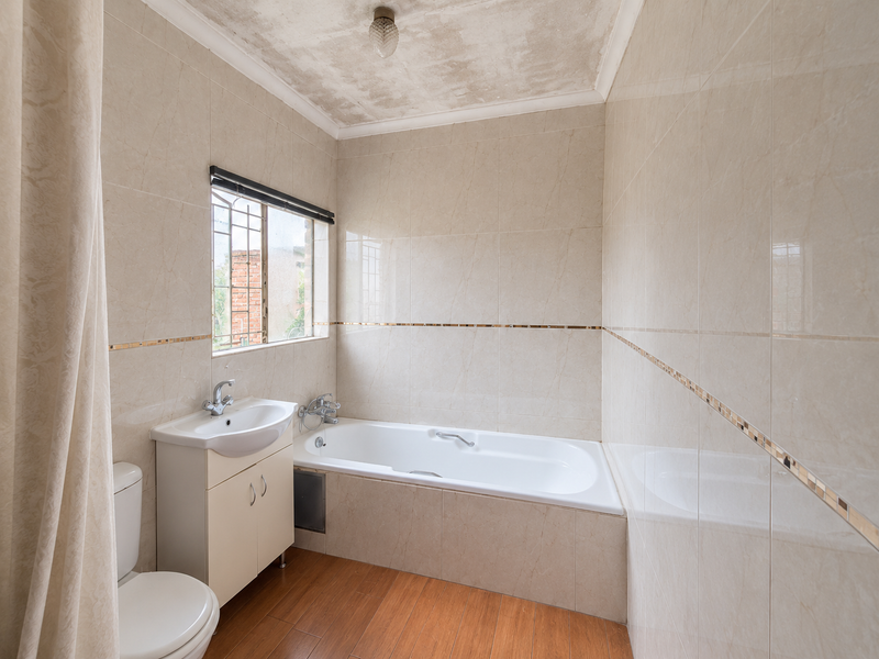 A functional bathroom featuring a built-in bathtub, vanity basin, and toilet, with tiled walls and wood-look flooring. The ceiling requires maintenance and repainting.