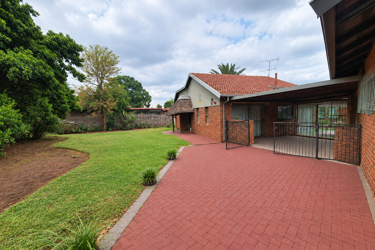 Rear view of the property featuring a spacious lawn, red brick paved patio area, and a covered outdoor space with security gate access.