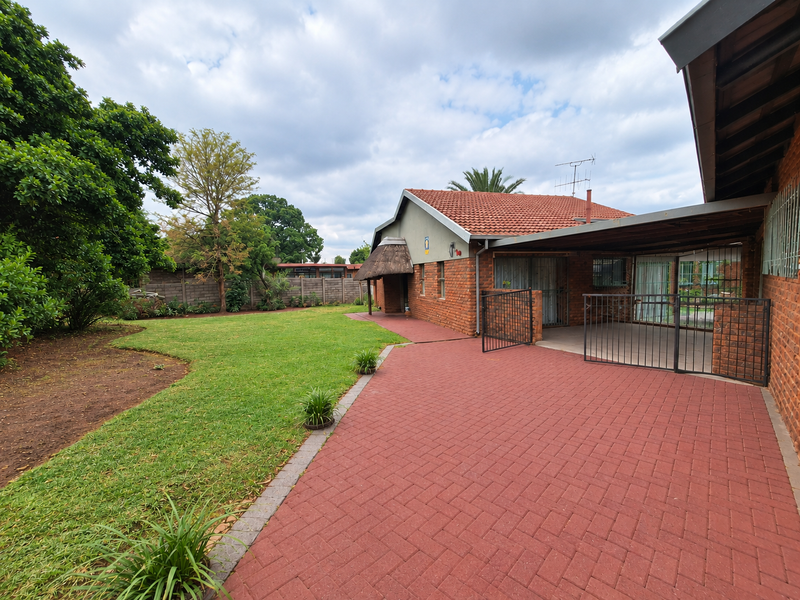 Rear view of the property featuring a spacious lawn, red brick paved patio area, and a covered outdoor space with security gate access.