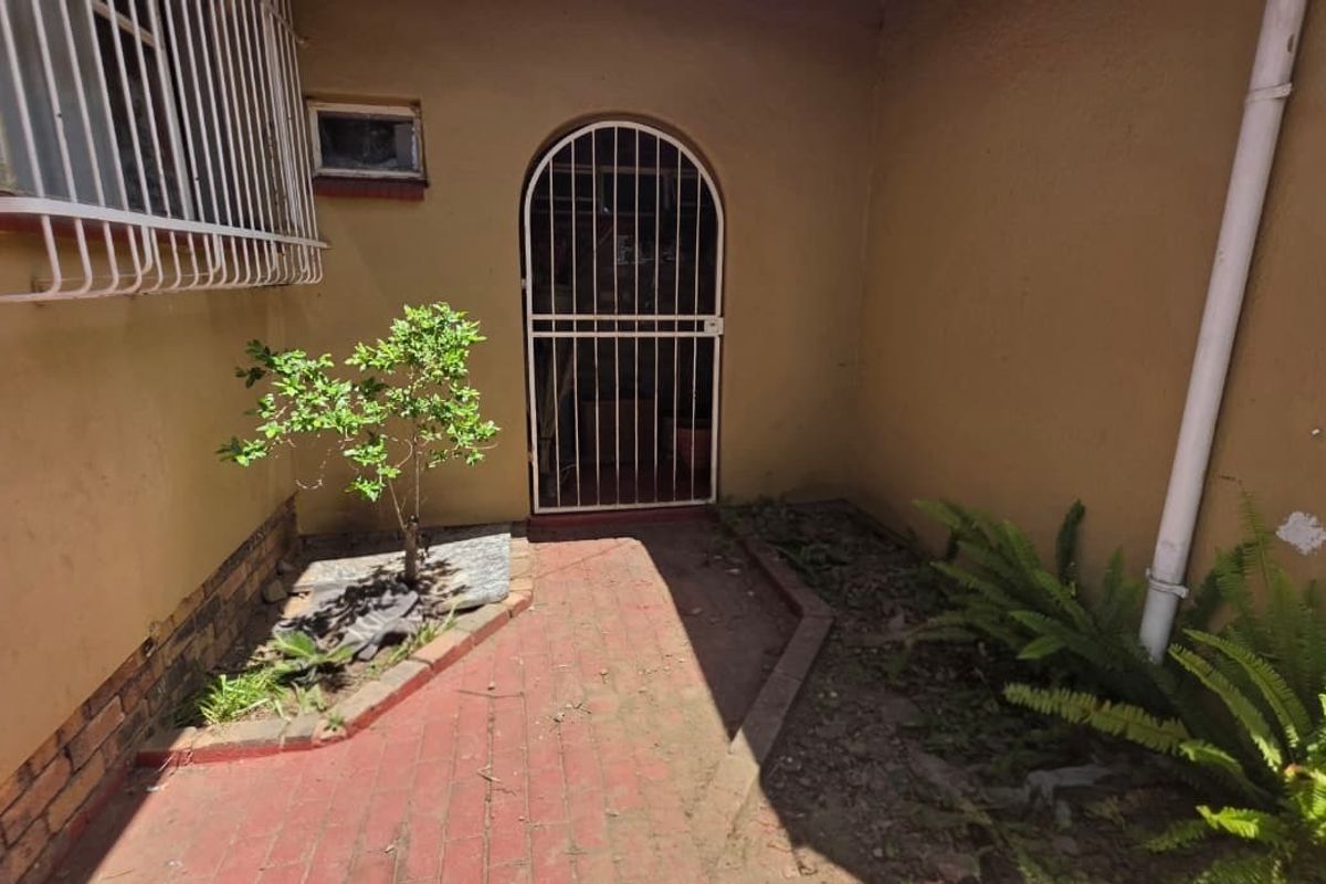 A rear exterior area featuring brick paving, a security gate, and security bars on the window, showing signs of wear and maintenance needs.