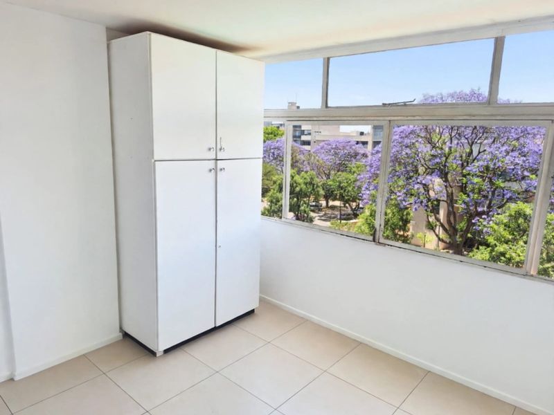 A bright, empty bedroom featuring tiled flooring, a built-in cupboard, and large windows offering a pleasant view of flowering trees.