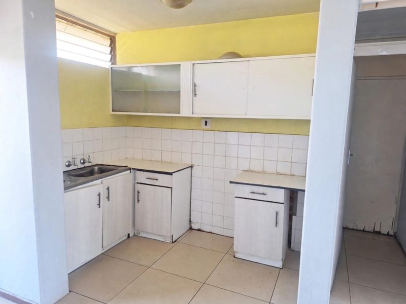 A dated kitchen featuring white built-in cabinetry, a stainless steel sink, tiled backsplash, and tiled flooring, offering significant potential for modernization.