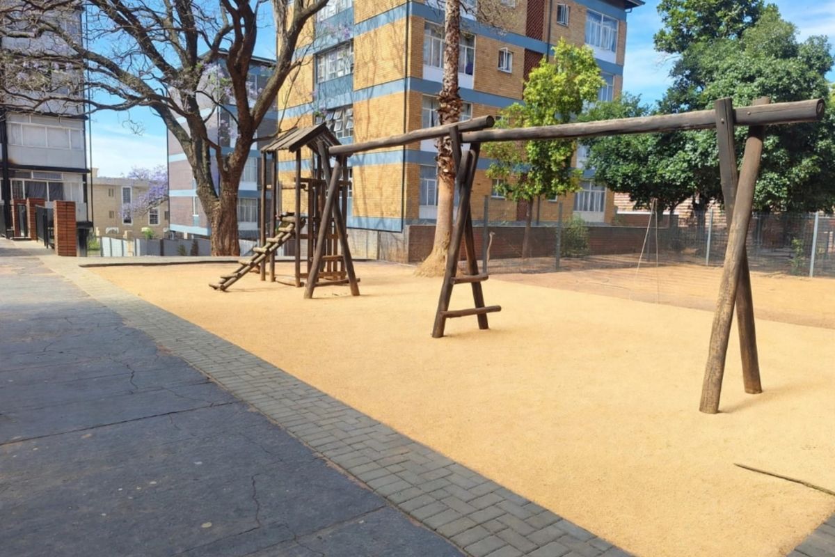 Communal outdoor area featuring a wooden playground structure set within a sand pit, bordered by paved walkways and residential apartment buildings.