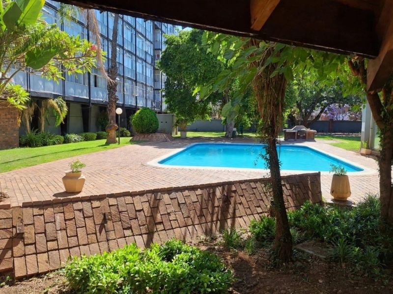 Communal pool area featuring a swimming pool, brick paved surround, and a built-in braai facility set within a landscaped garden.