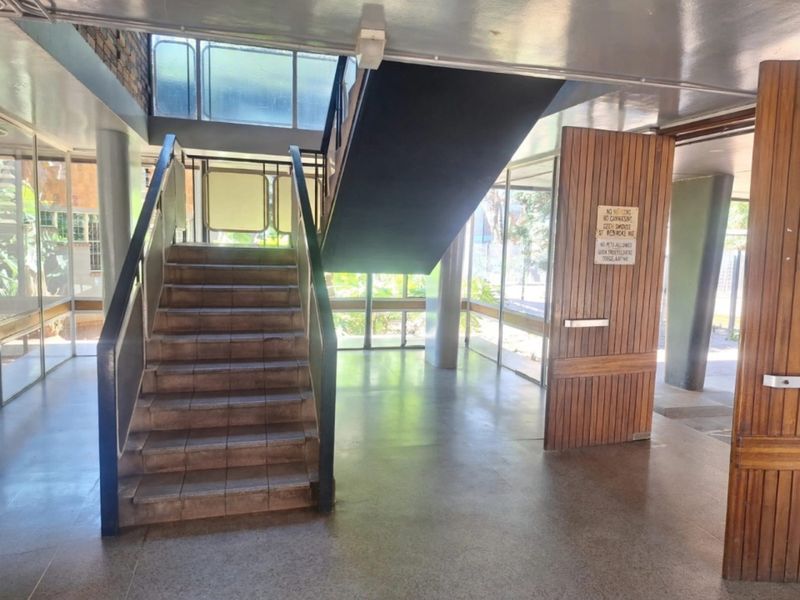 A spacious building entrance lobby featuring a central staircase, large glass windows, and durable terrazzo flooring with a prominent timber entrance door.
