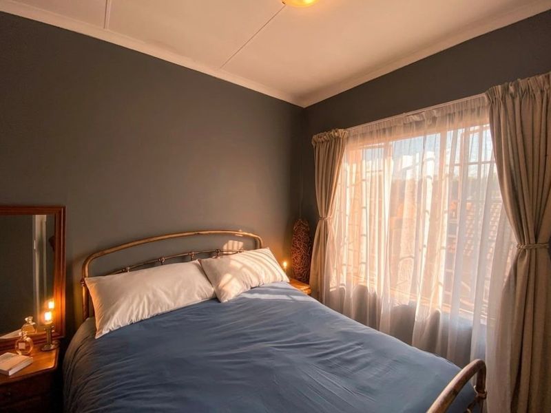 A cozy bedroom featuring dark painted walls, natural light through a window with security bars, and neutral curtains.