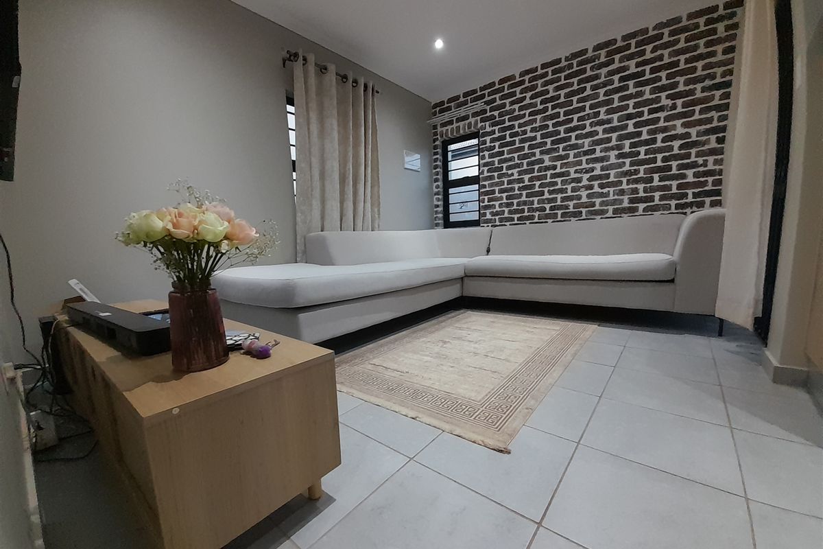 A comfortable living room featuring a distinctive exposed brick accent wall, neutral tiled flooring, and modern recessed lighting.