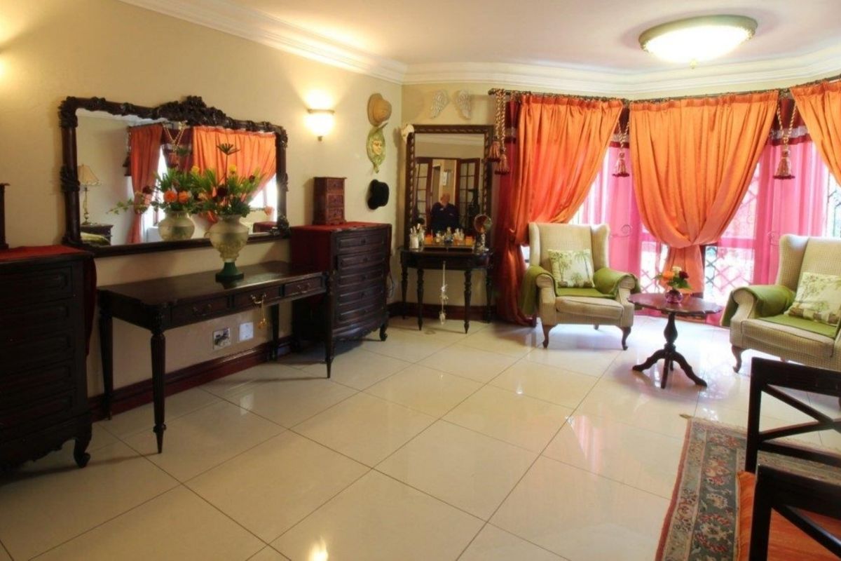 A spacious living room featuring polished tiled flooring, decorative cove cornicing, and wall-mounted lighting fixtures, offering a bright and well-maintained living space.