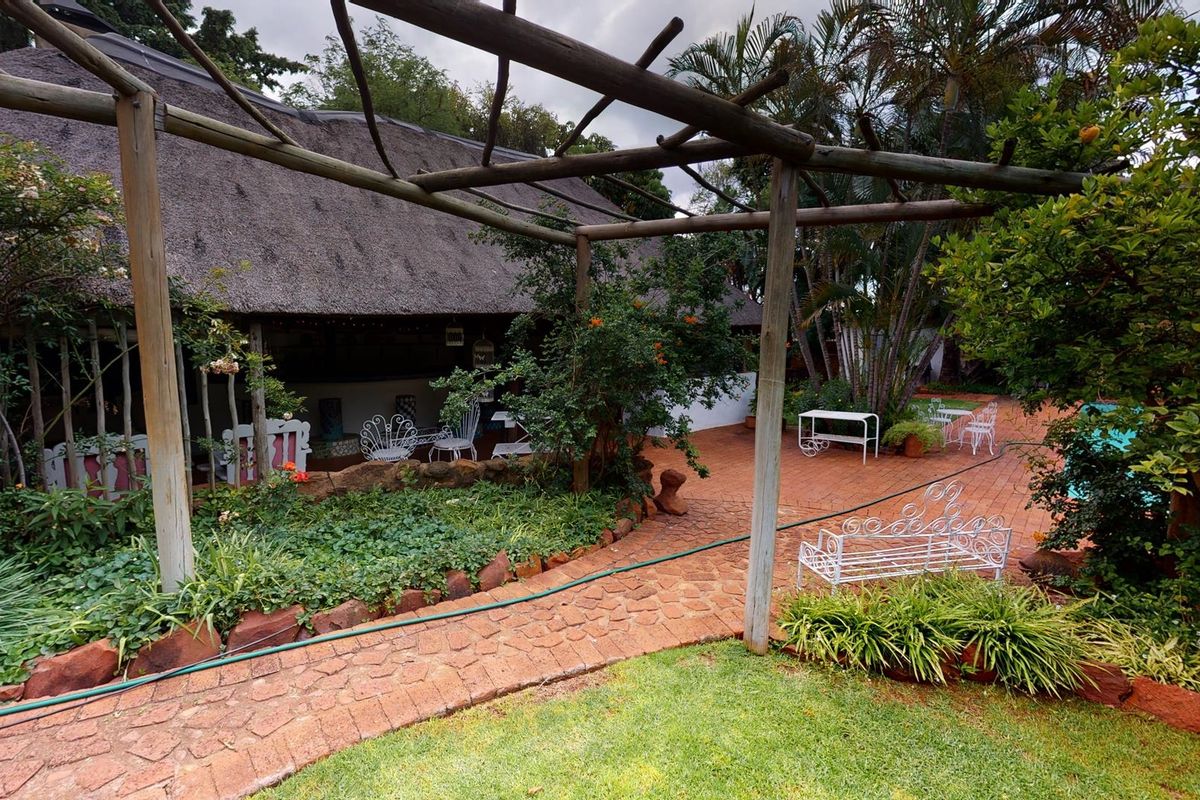 A rear garden view featuring a thatched roof dwelling, a timber pergola structure, brick paved pathways, and a swimming pool area.