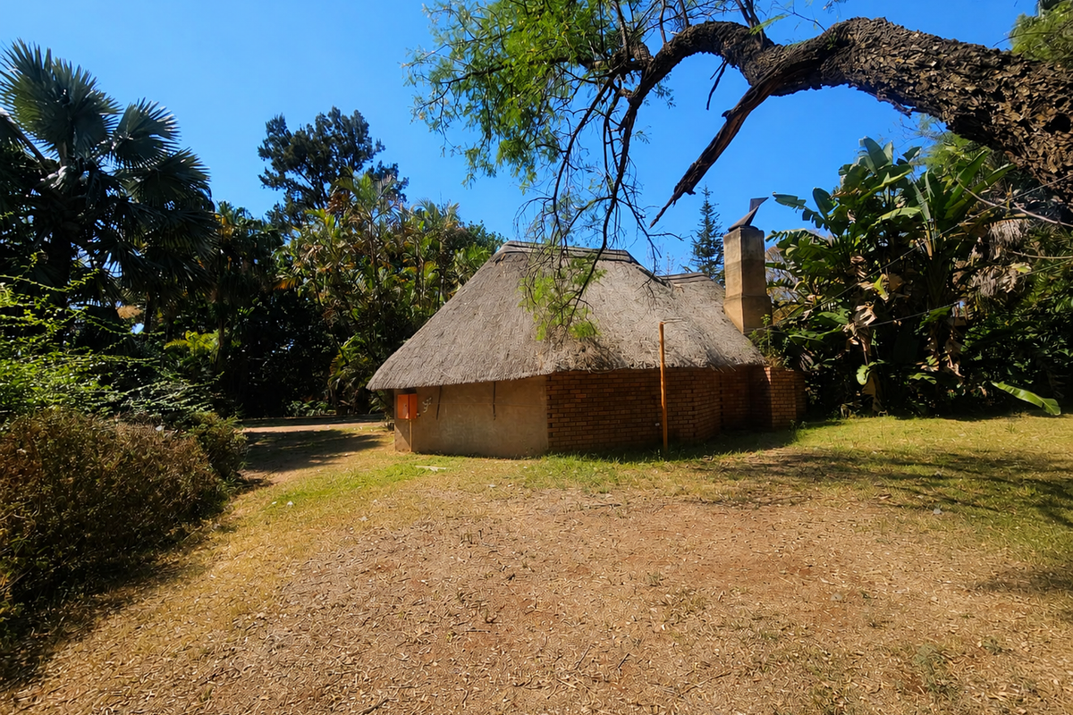 A view of a rustic thatched-roof structure with an external brick chimney, set within a large, mature garden area.