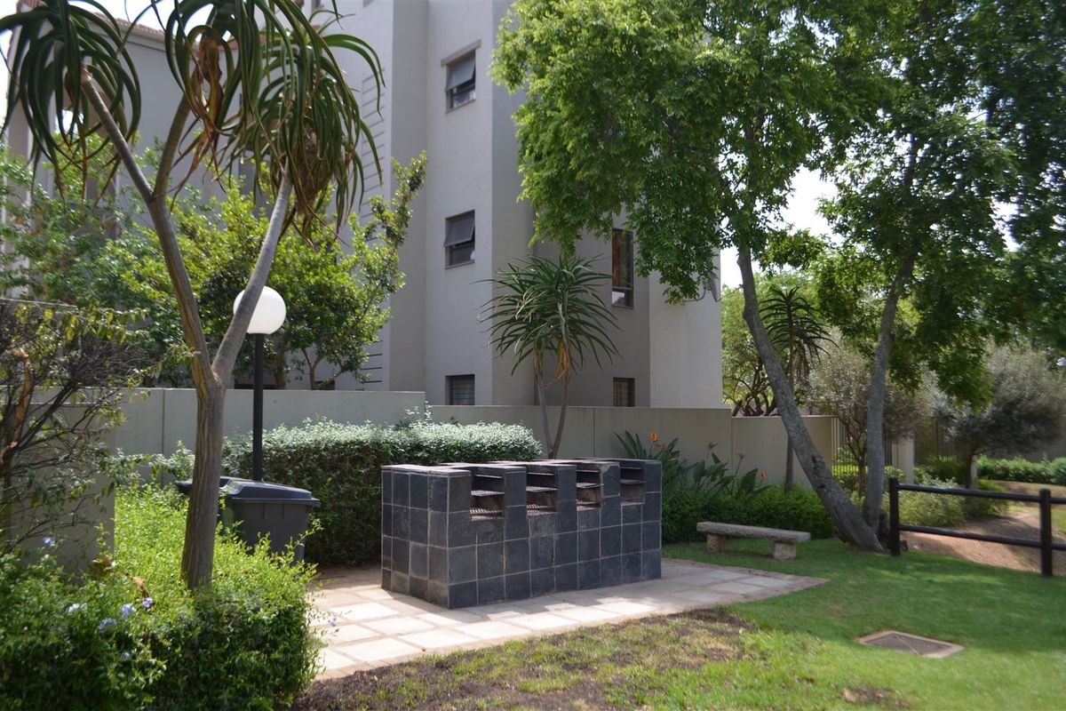 Communal outdoor area featuring a built-in tiled braai, paved patio space, and established garden surroundings within the apartment complex.