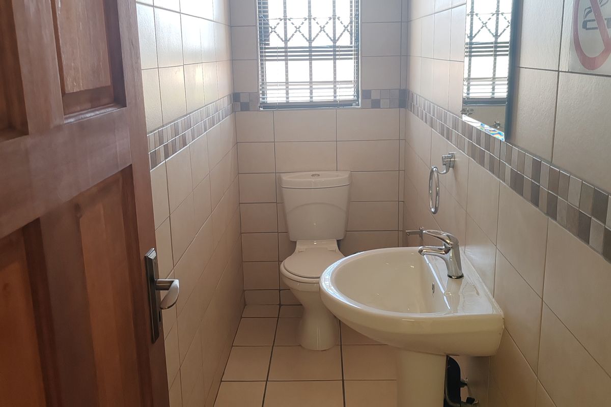 A compact commercial restroom featuring floor-to-ceiling wall tiles, a pedestal basin, a toilet, and a security-grilled window.