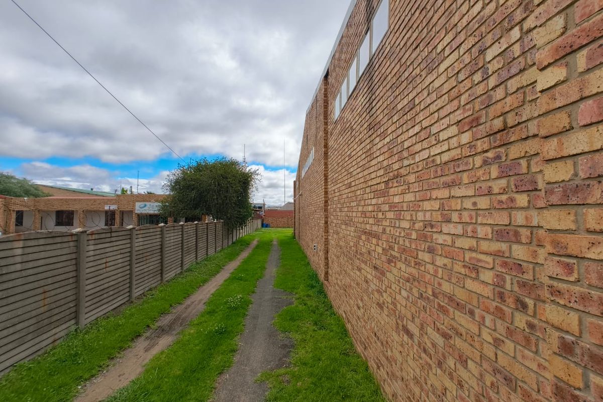 A side exterior view of the commercial property featuring a long brick facade and a narrow grass access path alongside a boundary fence.