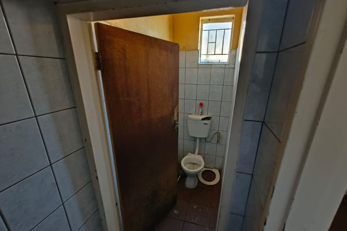 A small, dated restroom facility featuring a standard toilet, tiled walls, and a window with security bars, requiring significant maintenance and refurbishment.