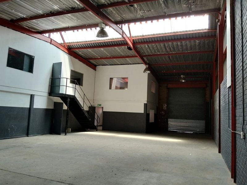 Spacious industrial warehouse unit featuring high ceilings with exposed steel trusses, a large roller shutter door, mezzanine level, and durable concrete flooring.