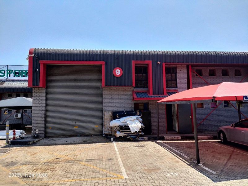 Industrial unit exterior featuring a large roller shutter door, brick facade, and dedicated paved parking with a shade cloth carport.