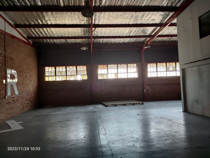 Spacious industrial warehouse unit featuring exposed brick walls, high steel roof trusses, concrete flooring, and large multi-pane windows providing natural light.