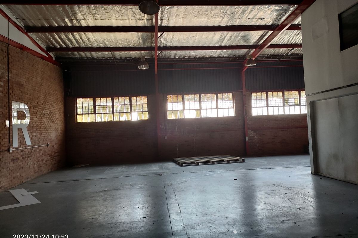 Spacious industrial warehouse unit featuring exposed brick walls, high steel roof trusses, concrete flooring, and large multi-pane windows providing natural light.