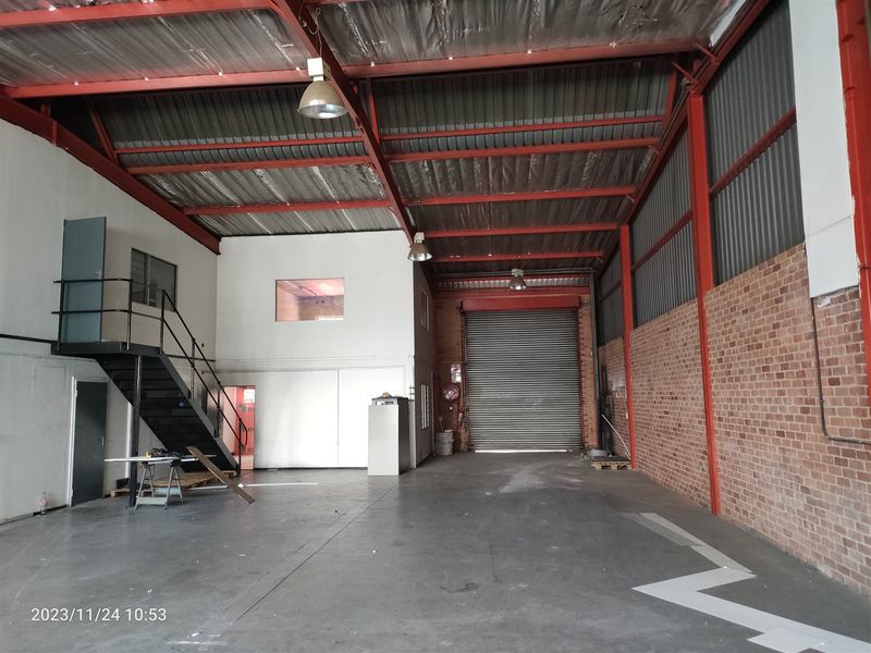 Spacious industrial warehouse unit featuring high ceilings with exposed steel trusses, a roller shutter door, mezzanine level, and durable concrete flooring.