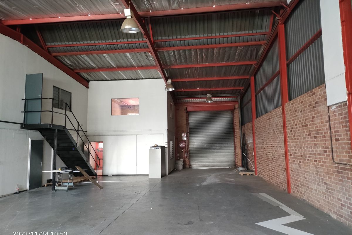Spacious industrial warehouse unit featuring high ceilings with exposed steel trusses, a roller shutter door, mezzanine level, and durable concrete flooring.