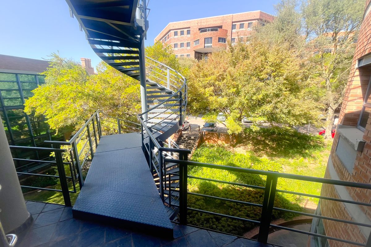 Exterior view featuring a metal spiral staircase providing access between levels, overlooking a landscaped garden area and office building surroundings.