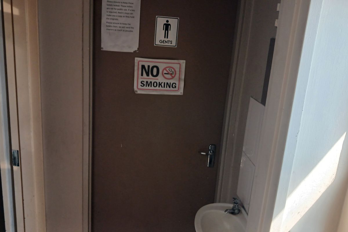 A functional gents restroom entrance featuring a wall-mounted basin and signage, showing signs of wear and dated finishes.