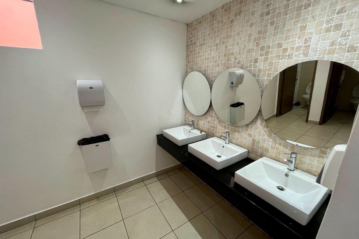 A clean, modern office restroom featuring a triple vanity with white ceramic basins, black countertop, and mosaic tiled wall.