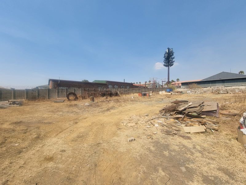 Vacant commercial land featuring a perimeter wall and a telecommunications mast, currently containing scattered debris and dry vegetation.