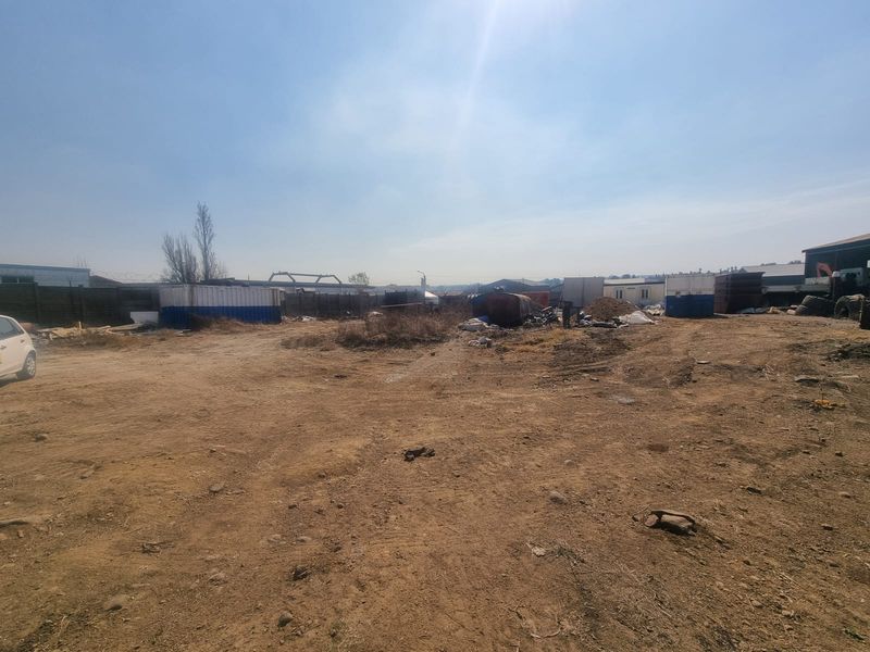 A wide view of a vacant commercial land plot featuring unpaved ground, perimeter fencing, and various storage containers on site.