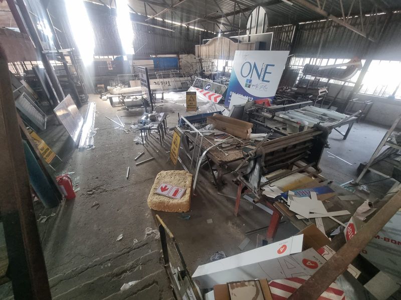 Large industrial warehouse space featuring exposed steel roof trusses, concrete flooring, and natural light from high-level windows, currently used for storage and manufacturing.