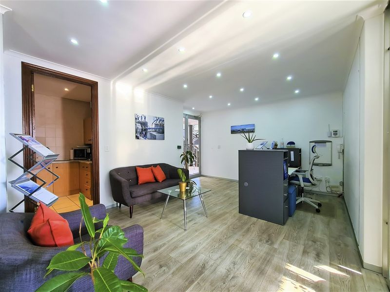 A well-lit professional office space featuring modern laminate flooring, recessed ceiling lighting, and an adjoining kitchenette area.