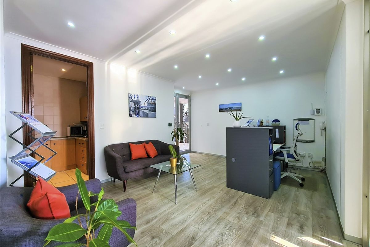 A well-lit professional office space featuring modern laminate flooring, recessed ceiling lighting, and an adjoining kitchenette area.