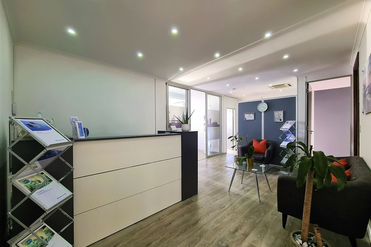 Modern office reception area featuring a built-in reception desk, recessed lighting, laminate flooring, and a glass partition leading to adjacent workspace.