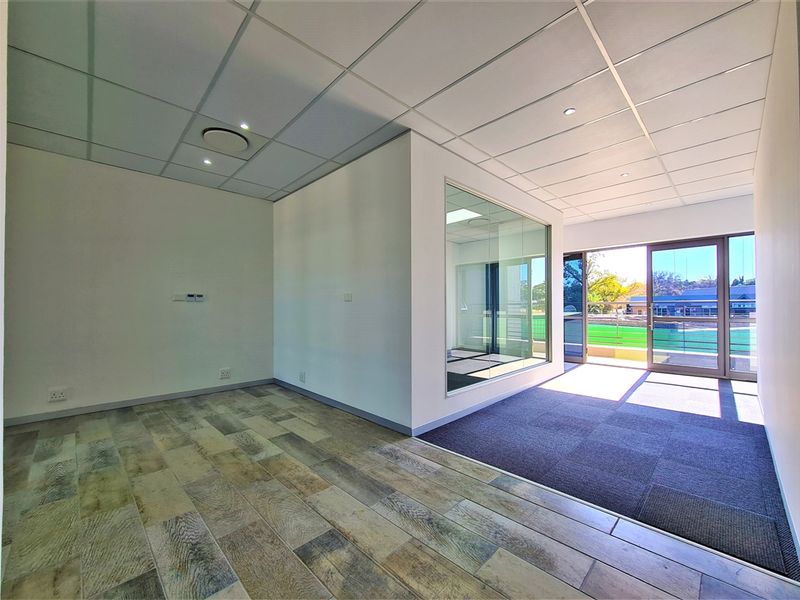 Modern, vacant office space featuring a glass partition, suspended ceiling with recessed lighting, and a mix of carpet and wood-look flooring with balcony access.