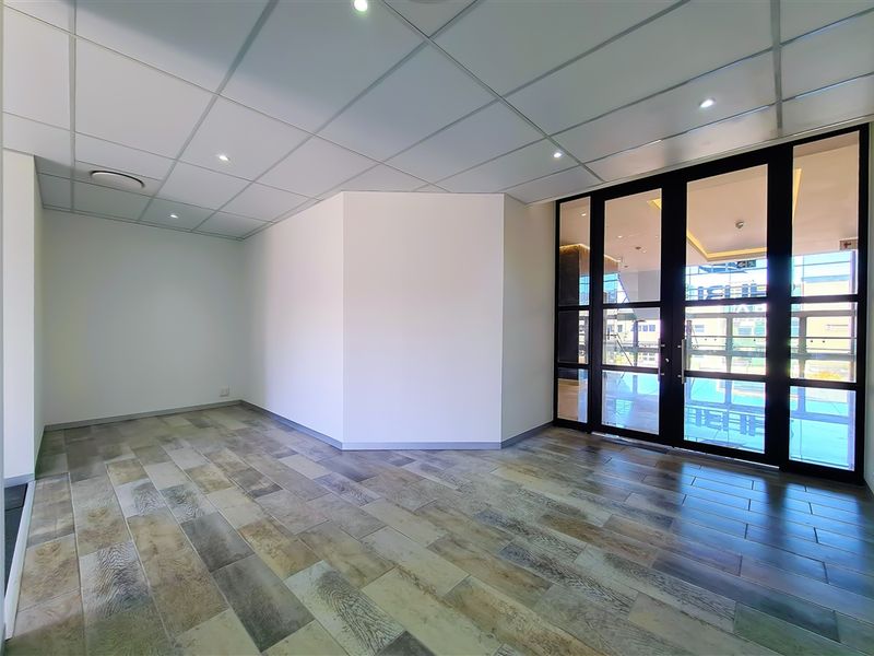 A bright, empty office space featuring modern wood-look tiled flooring, a suspended ceiling with recessed lighting, and large glass panel doors.