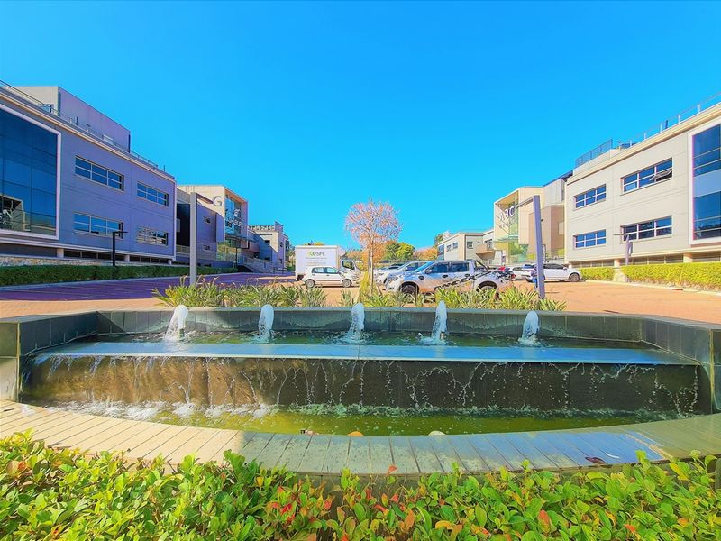 A modern office park entrance featuring a central water fountain, paved parking area, and contemporary multi-story office buildings with glass facades.