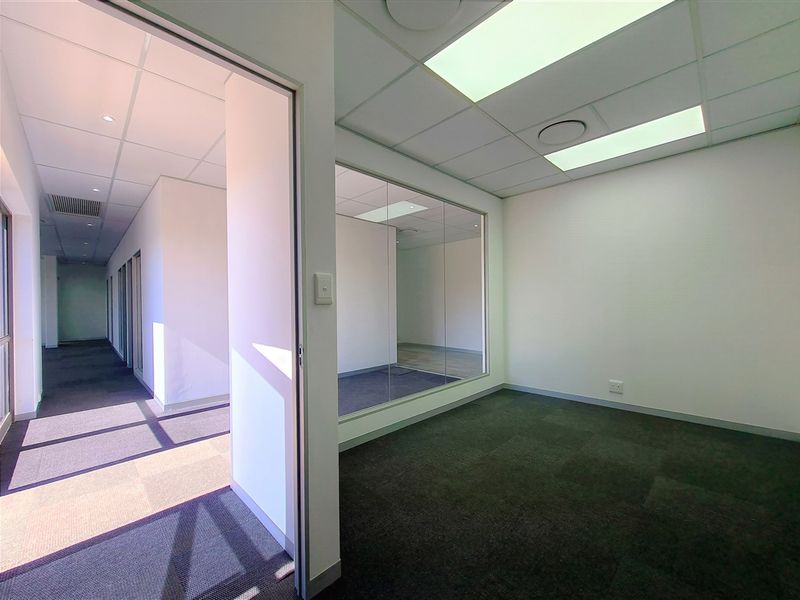 A clean, vacant office space featuring a glass partition wall, suspended ceiling with recessed lighting, and dark carpet tiles.