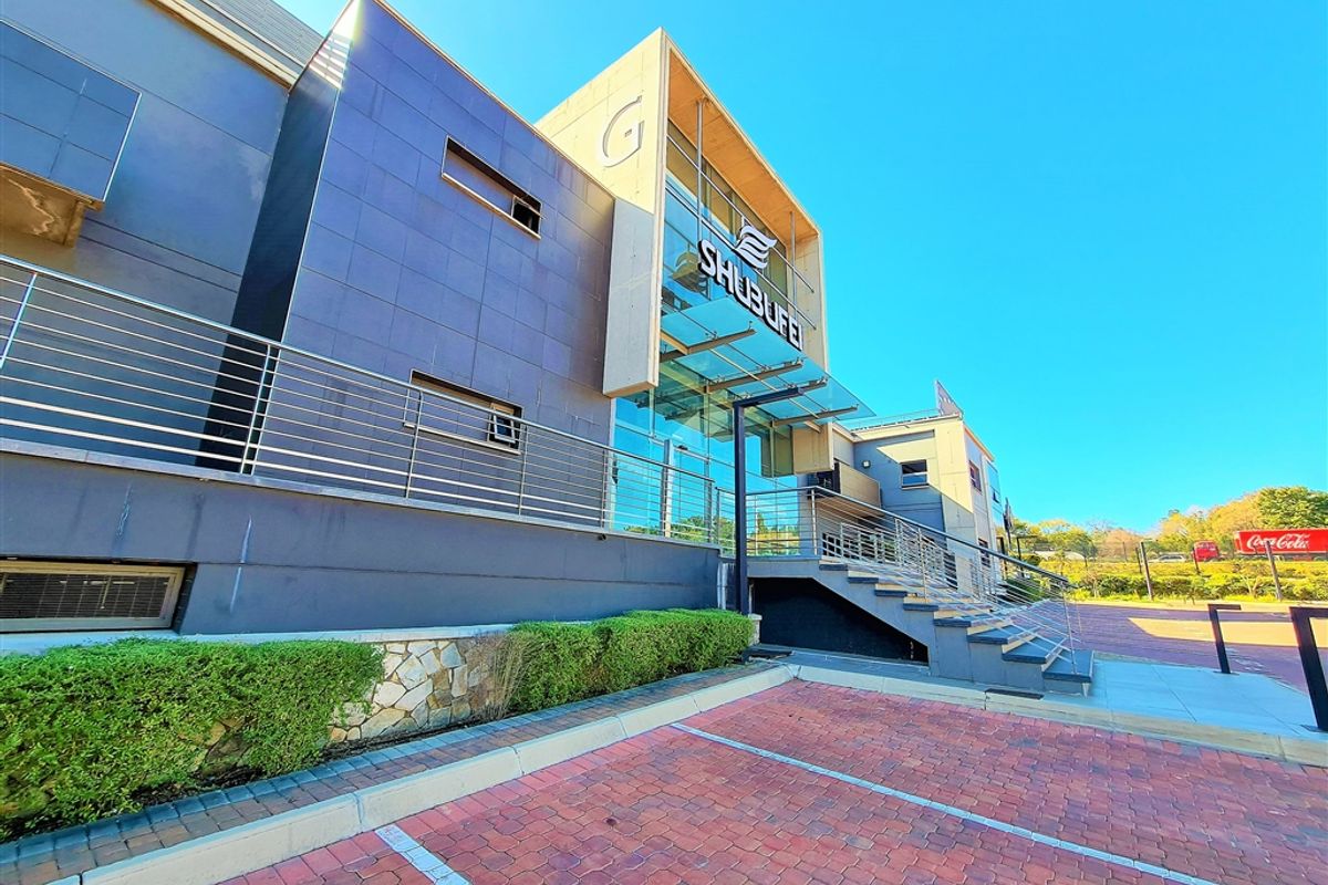 Modern office building exterior featuring a glass facade, stainless steel railings, and a paved parking area with landscaped hedging.