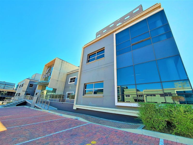Modern multi-story office building featuring a glass curtain wall facade, concrete architectural elements, and a paved parking area.