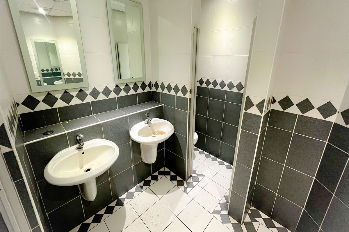 A functional office restroom featuring dual wall-mounted washbasins, tiled flooring, and partitioned toilet cubicles with a classic monochrome decorative border.