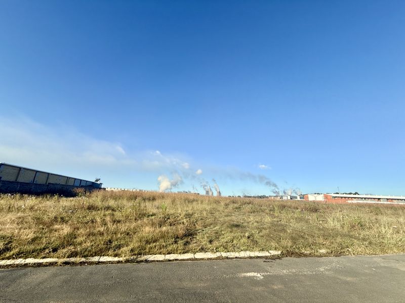 A view of a vacant commercial land plot featuring an asphalt road frontage and concrete curbing, with industrial buildings visible in the distance.
