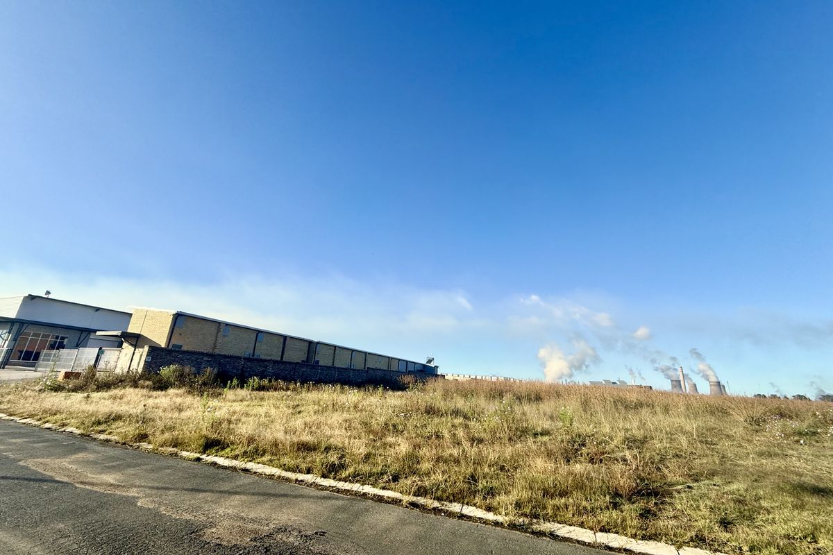 A view of a vacant commercial land plot featuring an asphalt road frontage, perimeter wall, and adjacent industrial infrastructure.