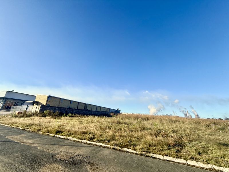 A view of a vacant commercial land plot featuring an asphalt road frontage, perimeter wall, and adjacent industrial infrastructure.