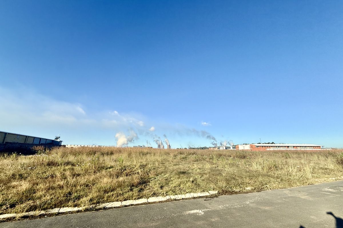 A view of a vacant commercial land plot featuring an adjacent asphalt road and concrete curbing, with industrial buildings visible in the distance.