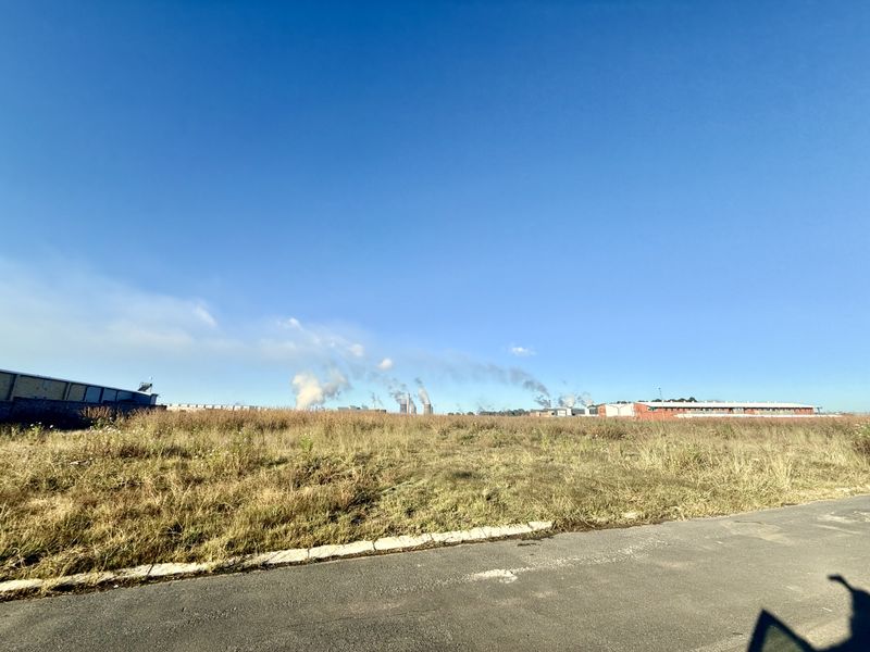 A view of a vacant commercial land plot featuring an adjacent asphalt road and concrete curbing, with industrial buildings visible in the distance.