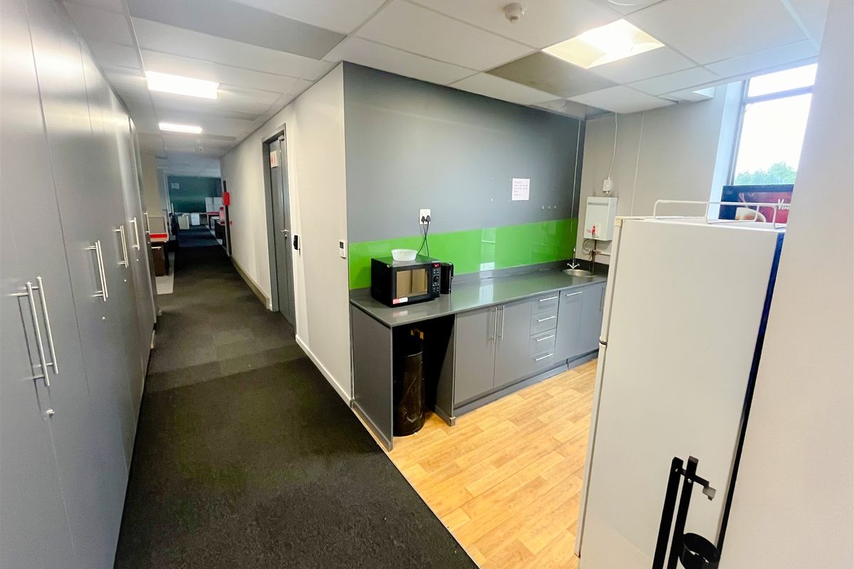 A functional office kitchenette area featuring built-in cabinetry, a sink, and a compact workspace, adjacent to a carpeted hallway.